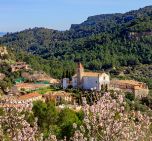 Bergdorf mit weißer Kirche, umgeben von Mandelblüten und grünen Hängen.