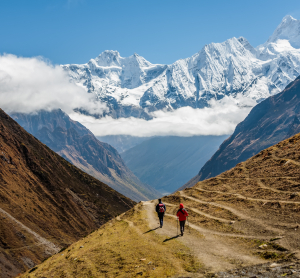 Zwei Wandernde auf einem Bergpfad zwischen steilen Tälern, vor schneebedeckten Himalaya-Gipfeln.