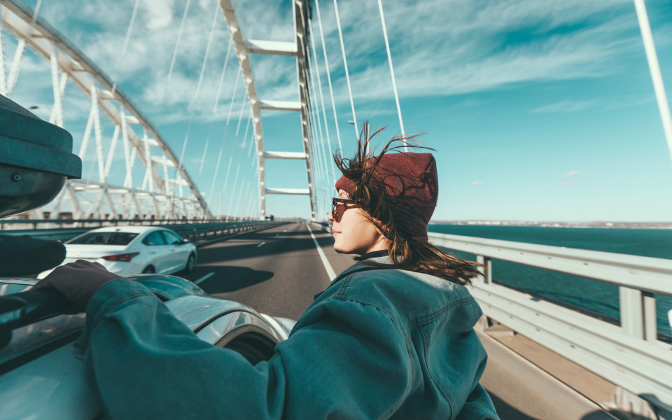 Person mit Mütze und Sonnenbrille lehnt sich aus einem fahrenden Auto auf einer Brücke, im Hintergrund fließt der Verkehr und das Meer ist sichtbar.