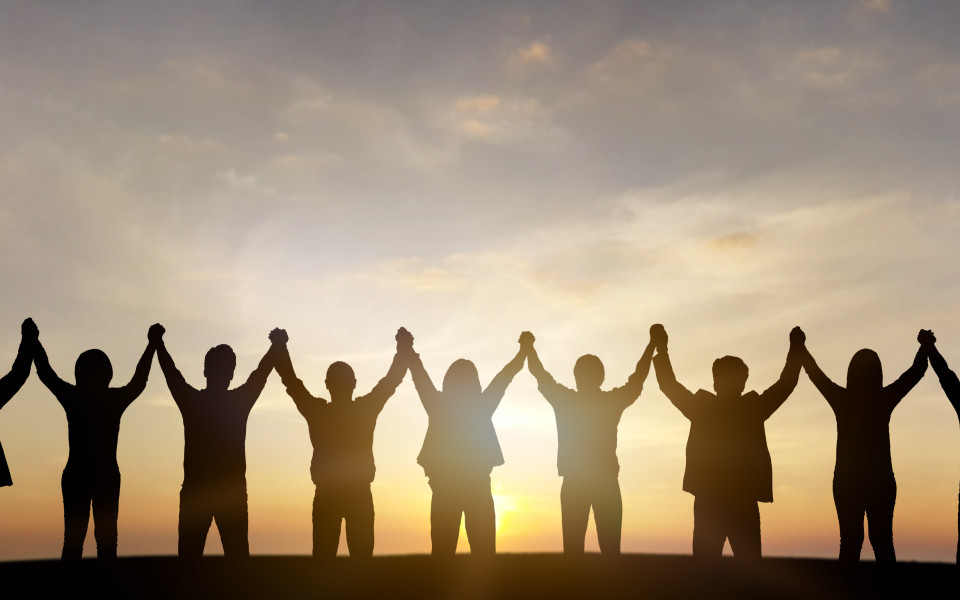 Silhouetten einer Gruppe von Menschen, die bei Sonnenuntergang nebeneinander stehen und die Hände in die Höhe halten.